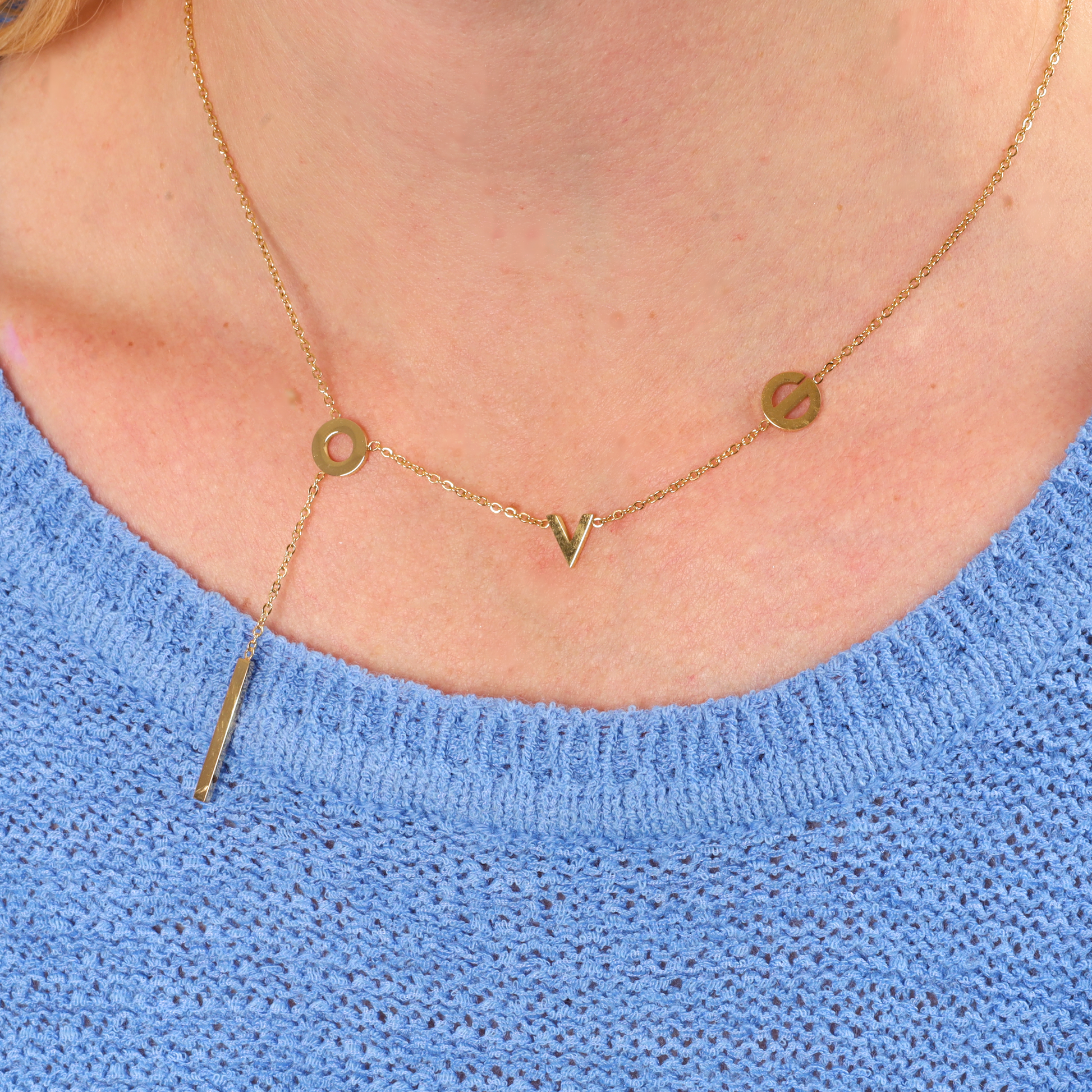 a water-resistant gold necklace with the letters "L", "O", "V", and "E" arranged along the chain. The necklace features a stylish and minimalistic design, with the letters spread out and a vertical bar pendant hanging elegantly below the "O". The necklace rests comfortably on a person wearing a blue knitted sweater, highlighting its versatility and modern appeal - Mia Ishaaq