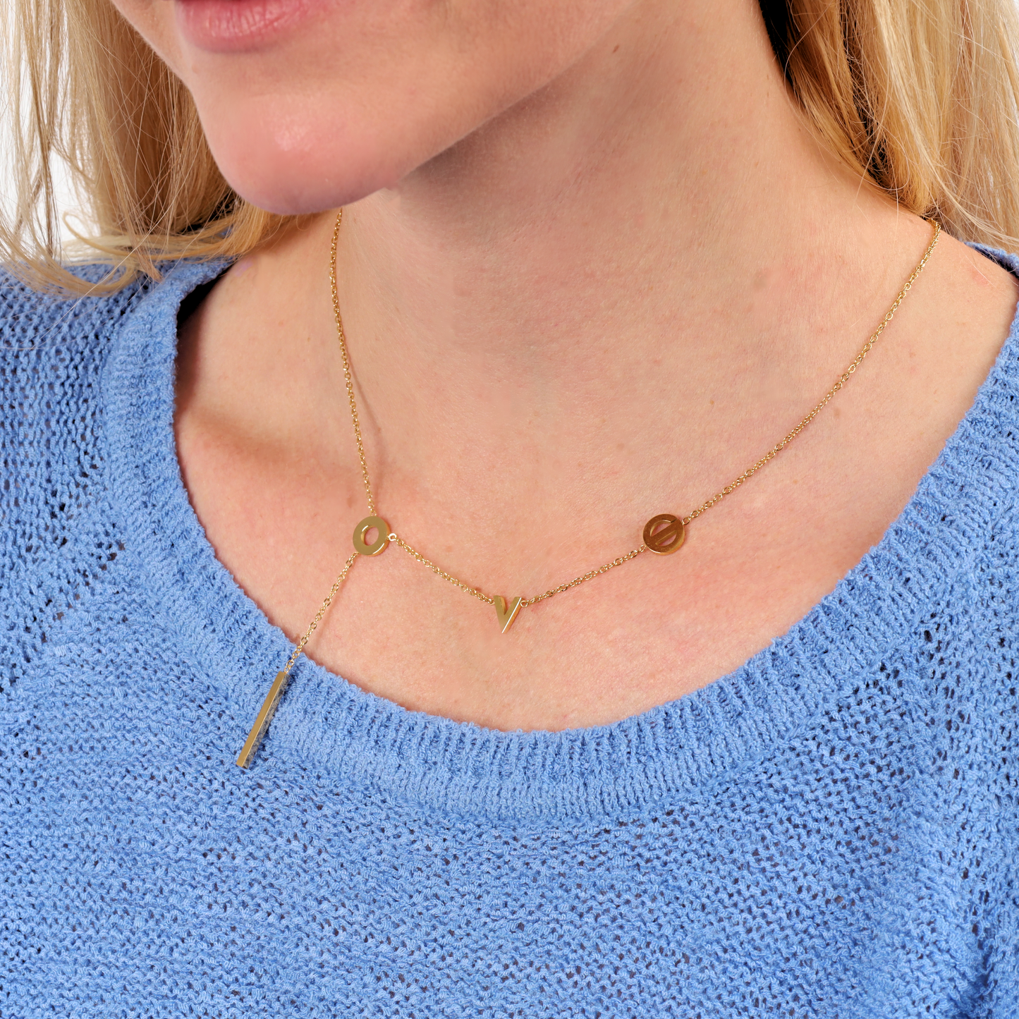 A close-up of a water-resistant gold necklace featuring the letters "L", "O", "V", and "E" spaced along a delicate chain. The necklace includes a sleek vertical bar pendant hanging below the letter "O", adding a modern touch. It rests elegantly on a person wearing a light blue knit sweater, showcasing its versatility and stylish, minimalist design - Mia Ishaaq