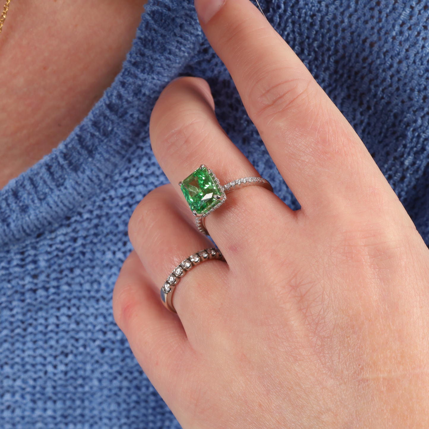 A hand showcasing Mia Ishaaq's Crushed Ice Green Radiant Promise Ring, featuring a sparkling green cubic zirconia stone and a slender diamond band, set against a blue knitted sweater backdrop.