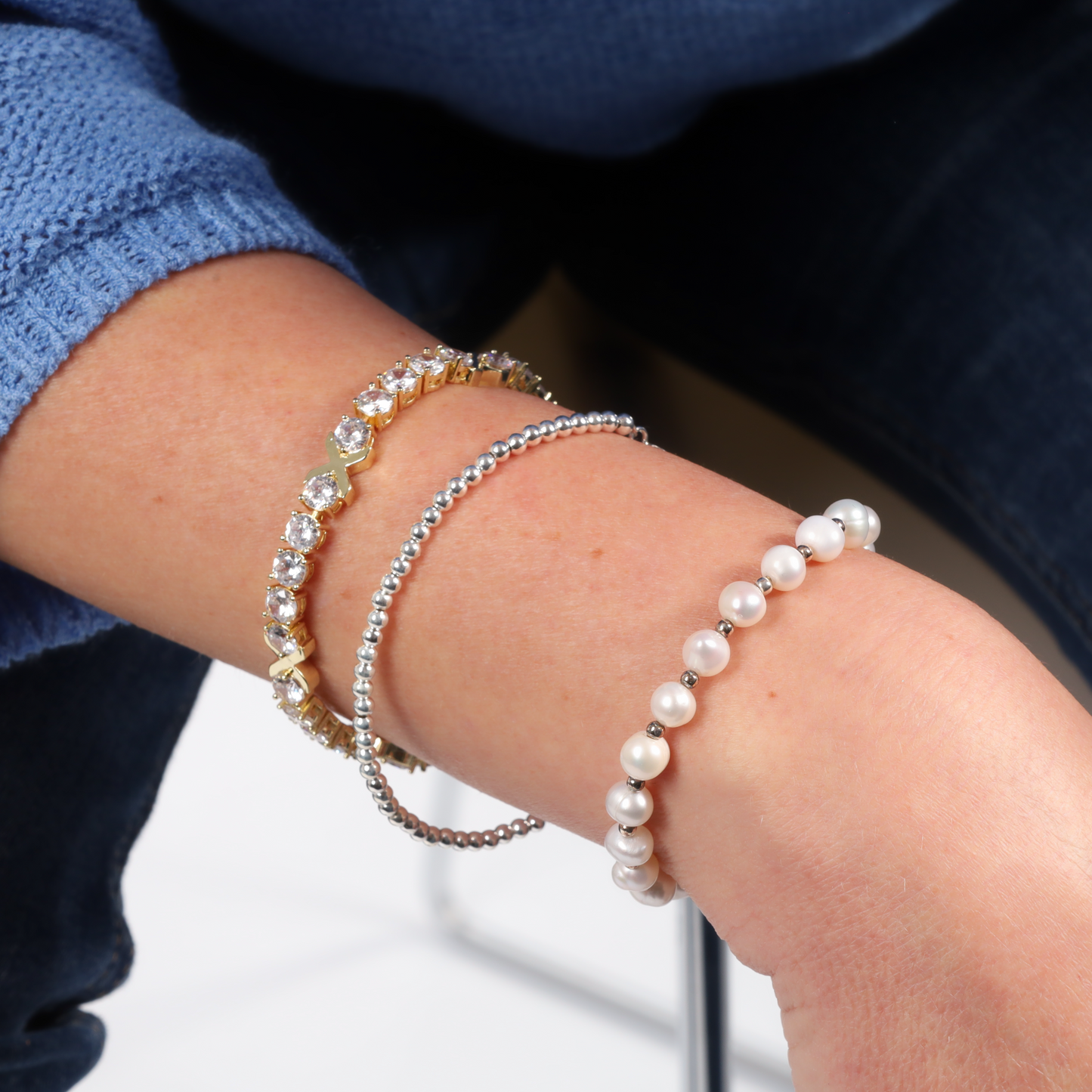 A person wearing three bracelets—a gold with stones, a silver beaded, and a pearl one over a blue sleeve— all complemented by the shimmer of the Mia Ishaaq Gemma XO Cross Love Bracelet in Gold.