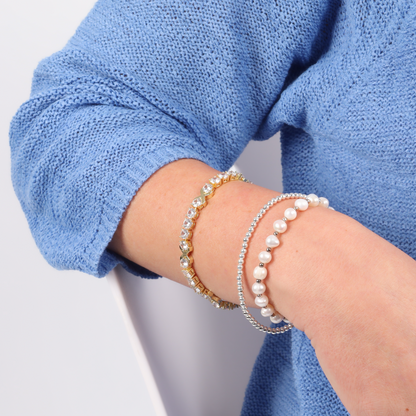 A close-up reveals a person wearing a blue sweater, their forearm elegantly adorned with pearl and gold-plated bracelets alongside the stunning Gemma XO Cross Love Bracelet in Gold by Mia Ishaaq.