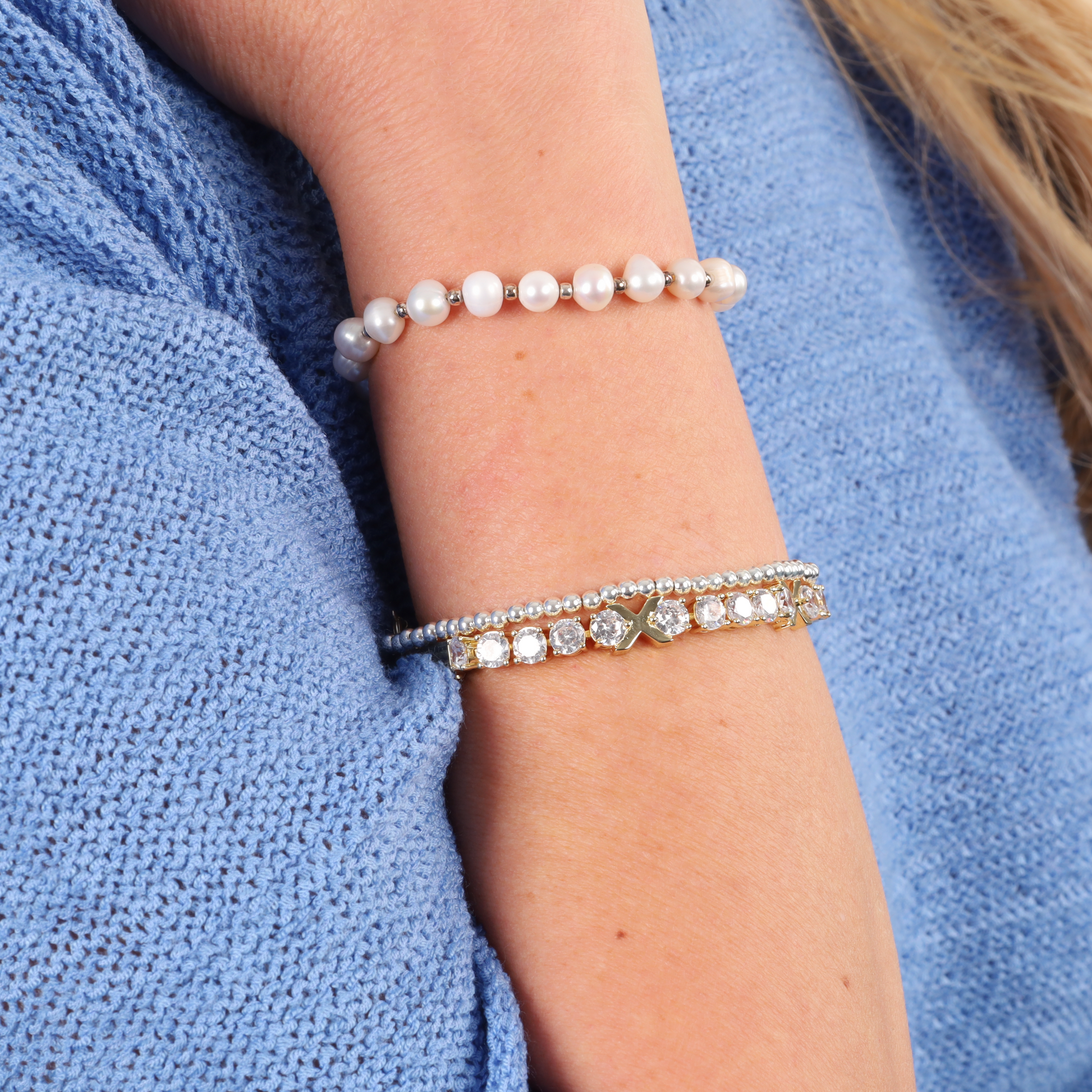 A close-up shows a woman's arm adorned with pearl and jeweled bracelets, featuring the stunning Gemma XO Cross Love Bracelet by Mia Ishaaq. The bracelet's dazzling gold plating and Cubic Zirconia accents perfectly contrast with the blue textured sweater in the background.