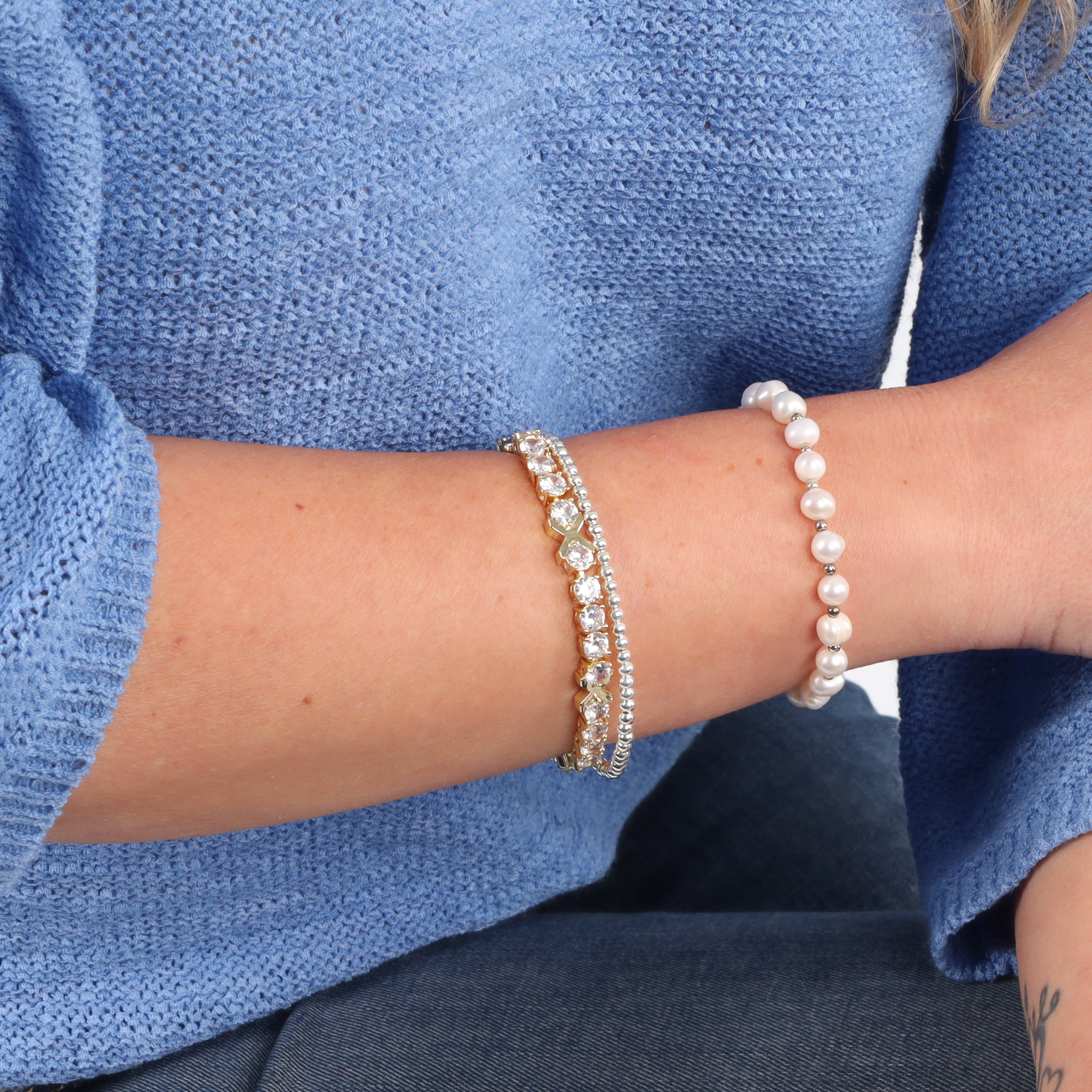 A woman in a blue sweater showcases her elegant jewelry collection, featuring the Mia Ishaaq Gemma XO Cross Love Bracelet in gold, adorned with cubic zirconia, a silver piece shimmering with diamonds, and delicate white pearls.