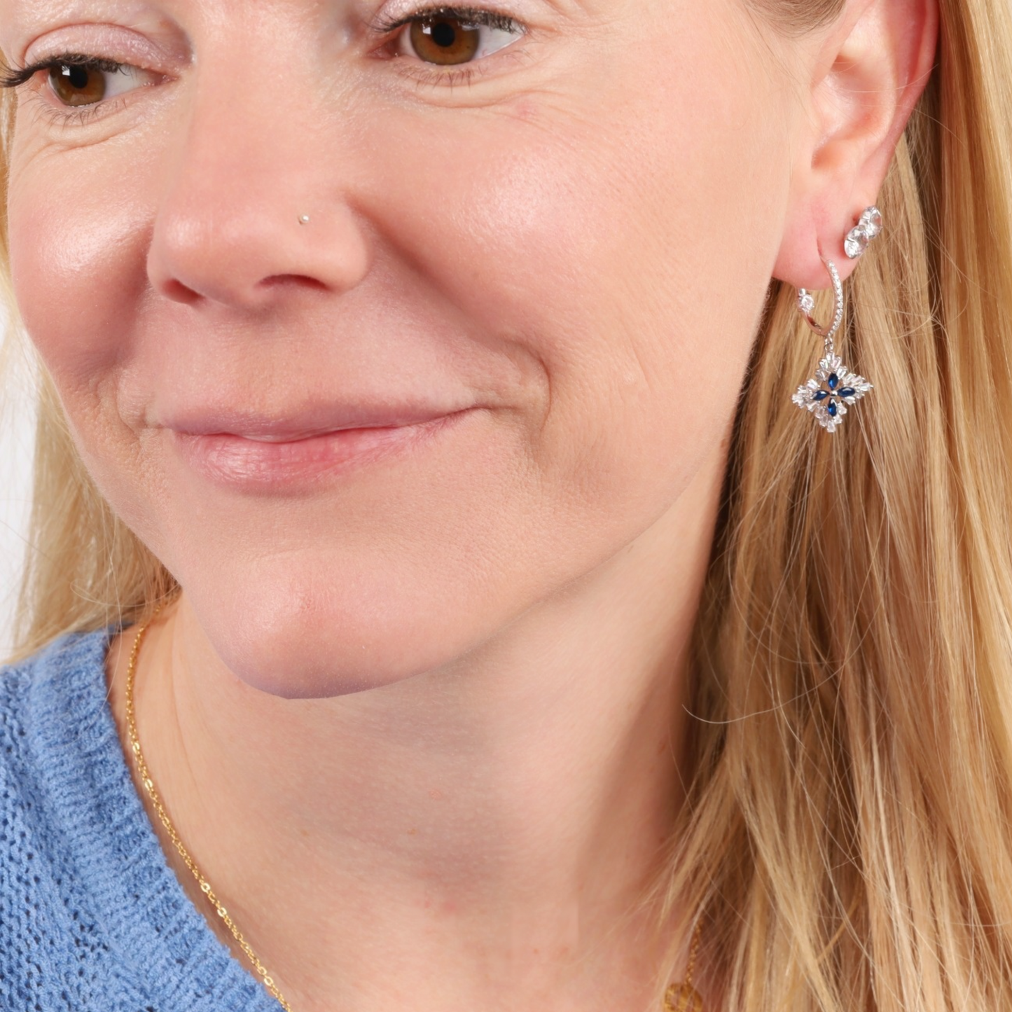 A woman with long blonde hair gently smiles while wearing the Blue Spinel CZ Earrings by Mia Ishaaq, crafted from 925 Sterling Silver, paired with a blue sweater.