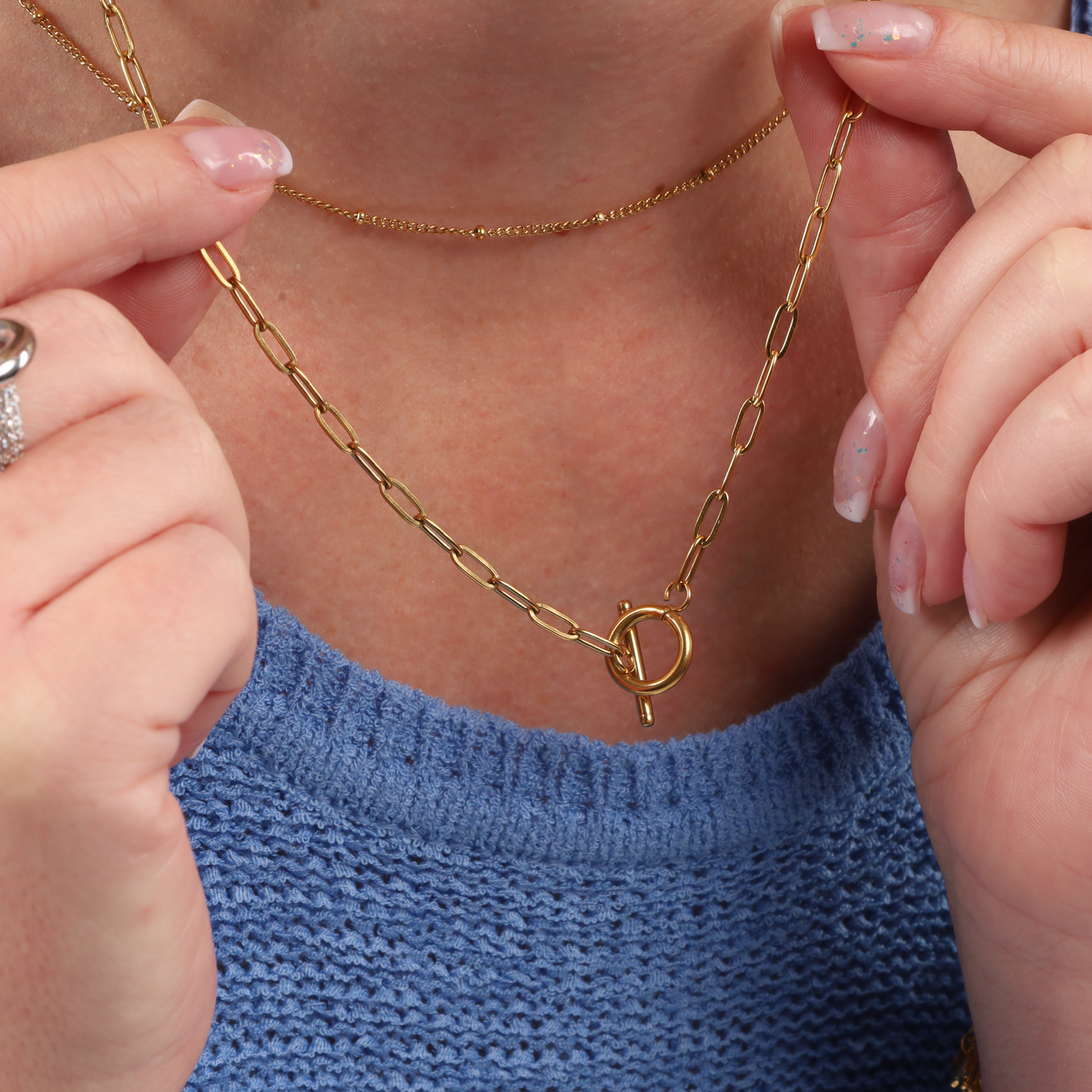 A person wearing the Mia Ishaaq Link Chain T-Bar Layering Necklace, which includes two gold link chain necklaces—one adorned with a toggle clasp and showcasing 18k gold plating—set against a blue knitted top.