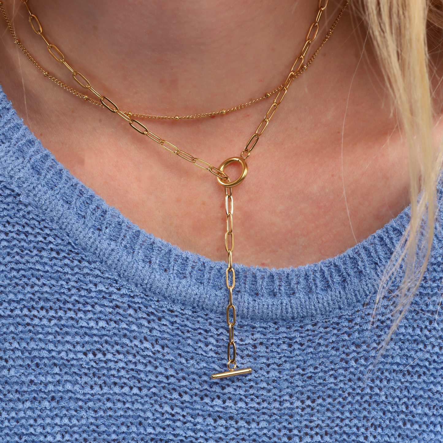 A person wears a blue knitted sweater adorned with Mia Ishaaq's Link Chain T-Bar Layering Necklace, effortlessly blending elegance with style.