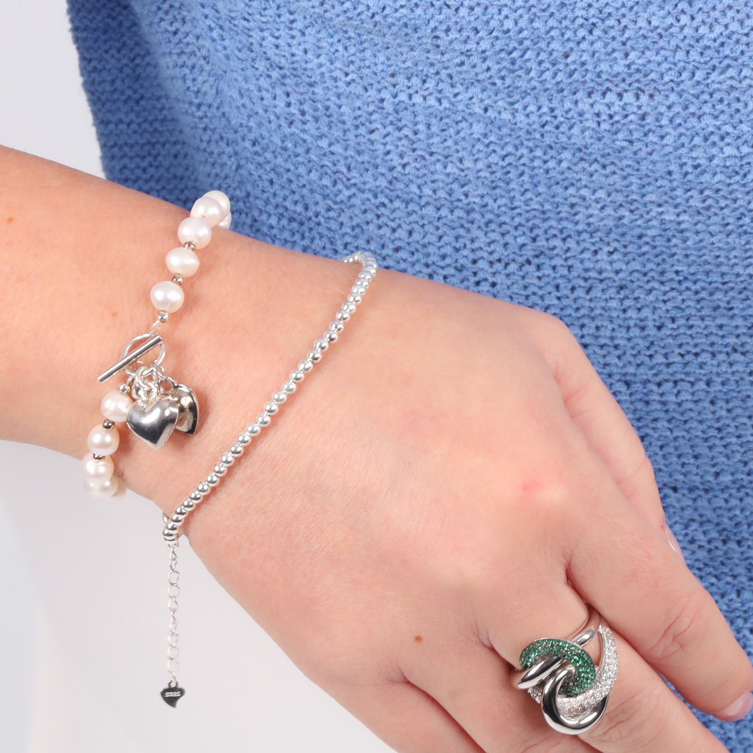 A hand adorned with the Mia Ishaaq Sterling Silver Beaded Bracelet and a ring featuring a green gemstone rests gracefully against a blue fabric background.