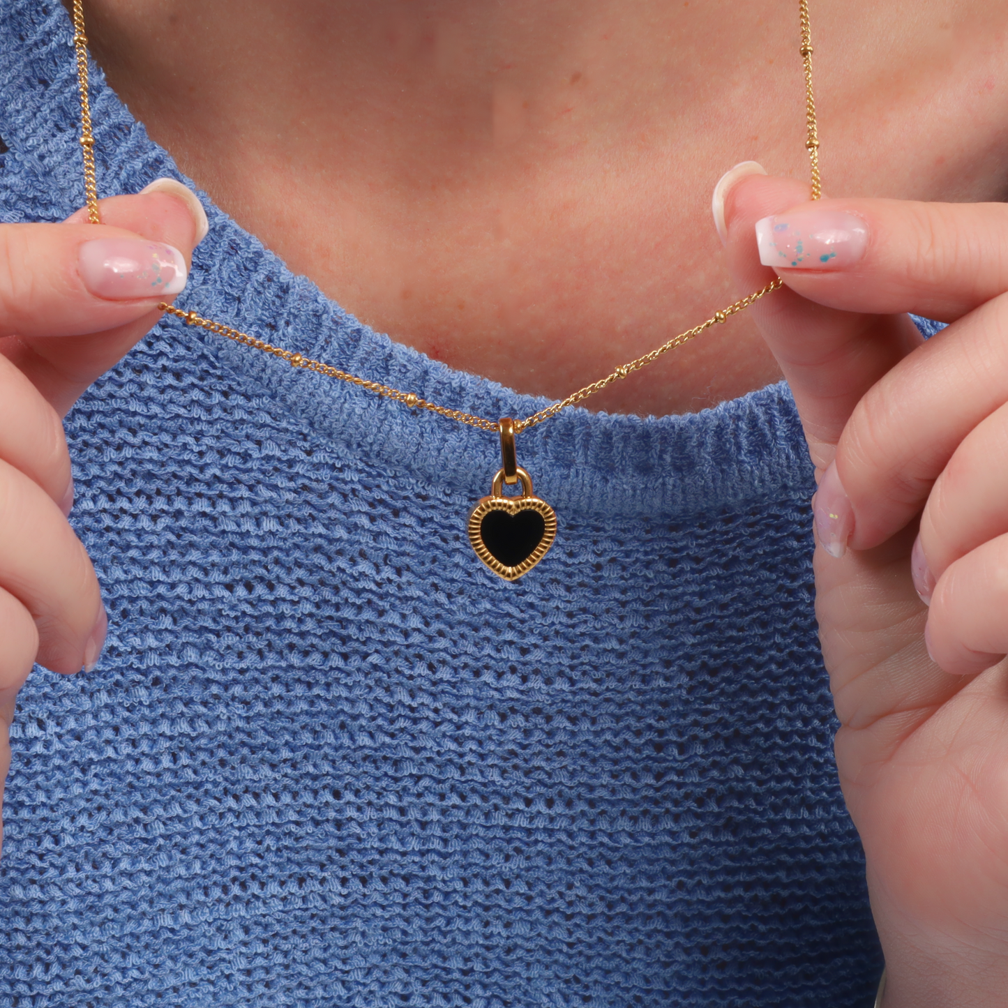 A person holds the Enamel Heart Reversible Necklace by Mia Ishaaq®, featuring an 18k gold reversible heart pendant, while wearing a blue textured top.