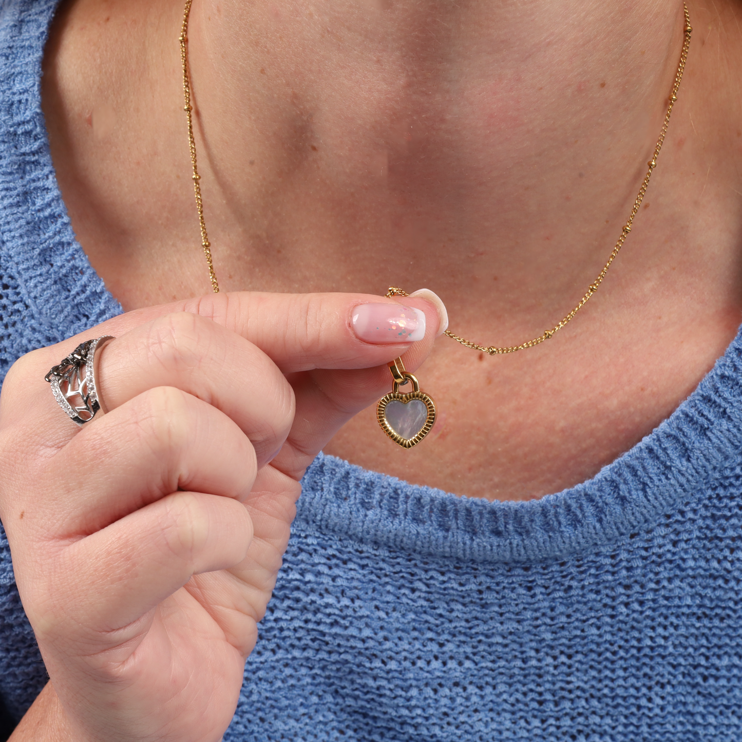 A person elegantly showcases the Mia Ishaaq® Enamel Heart Reversible Necklace, crafted in 18k gold, set against the cozy texture of a blue sweater.