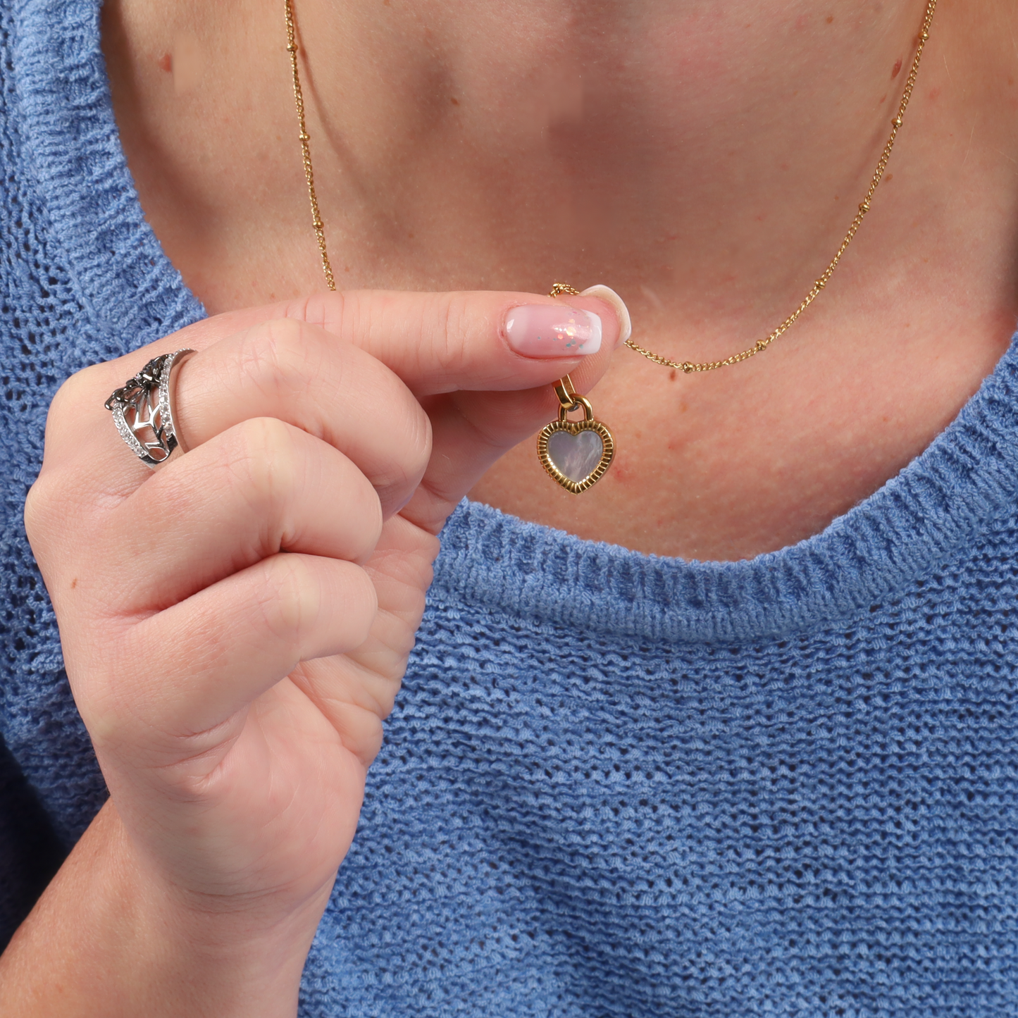 A woman holds the Enamel Heart Reversible Necklace by Mia Ishaaq® next to her knitted blue sweater, showcasing her versatile style. She's wearing a silver ring, perfectly complementing the elegant piece.