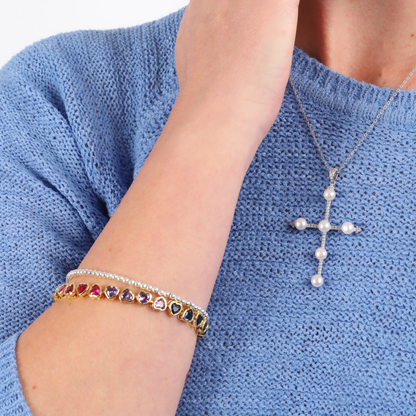 The person is dressed in a blue sweater, complemented by Mia Ishaaq's Rainbow Heart Tennis Bracelet featuring multi-colored stones and a pearl cross necklace.