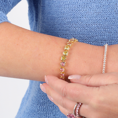 A person wearing the Rainbow Heart Tennis Bracelet by Mia Ishaaq featuring multi-colored stones, along with a blue knit sleeve.