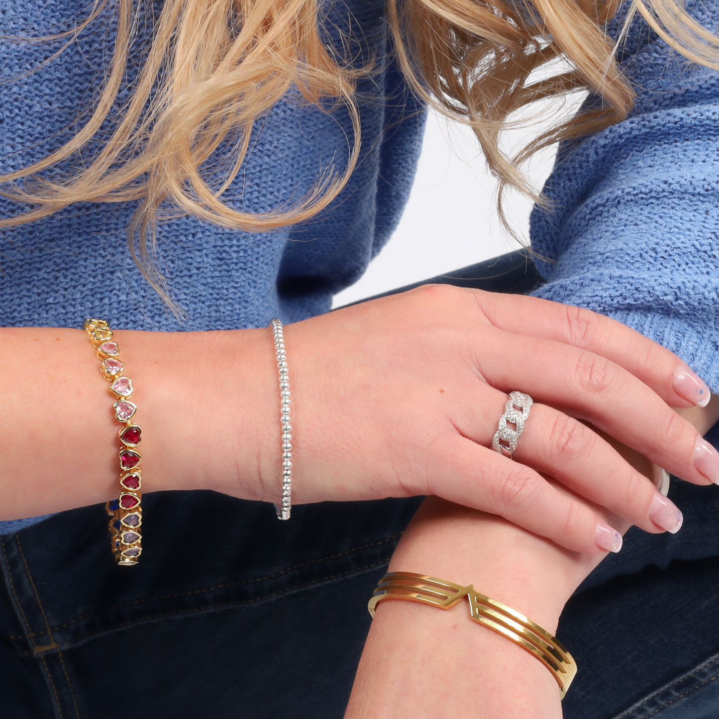 A person with long blonde hair and a blue sweater is wearing multiple gold and silver bracelets along with a ring, featuring the elegant Rainbow Heart Tennis Bracelet by Mia Ishaaq, which is adorned with multi-colored stones.