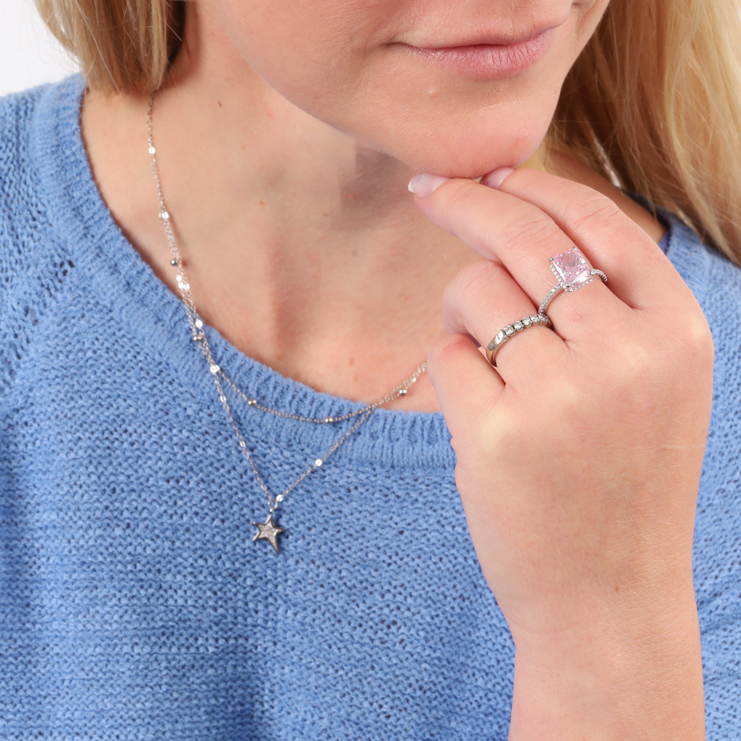 A woman in a blue sweater elegantly wears the Star Pendant Layered Celestial Necklace by Mia Ishaaq, showcasing a pink square-cut ring on her finger.