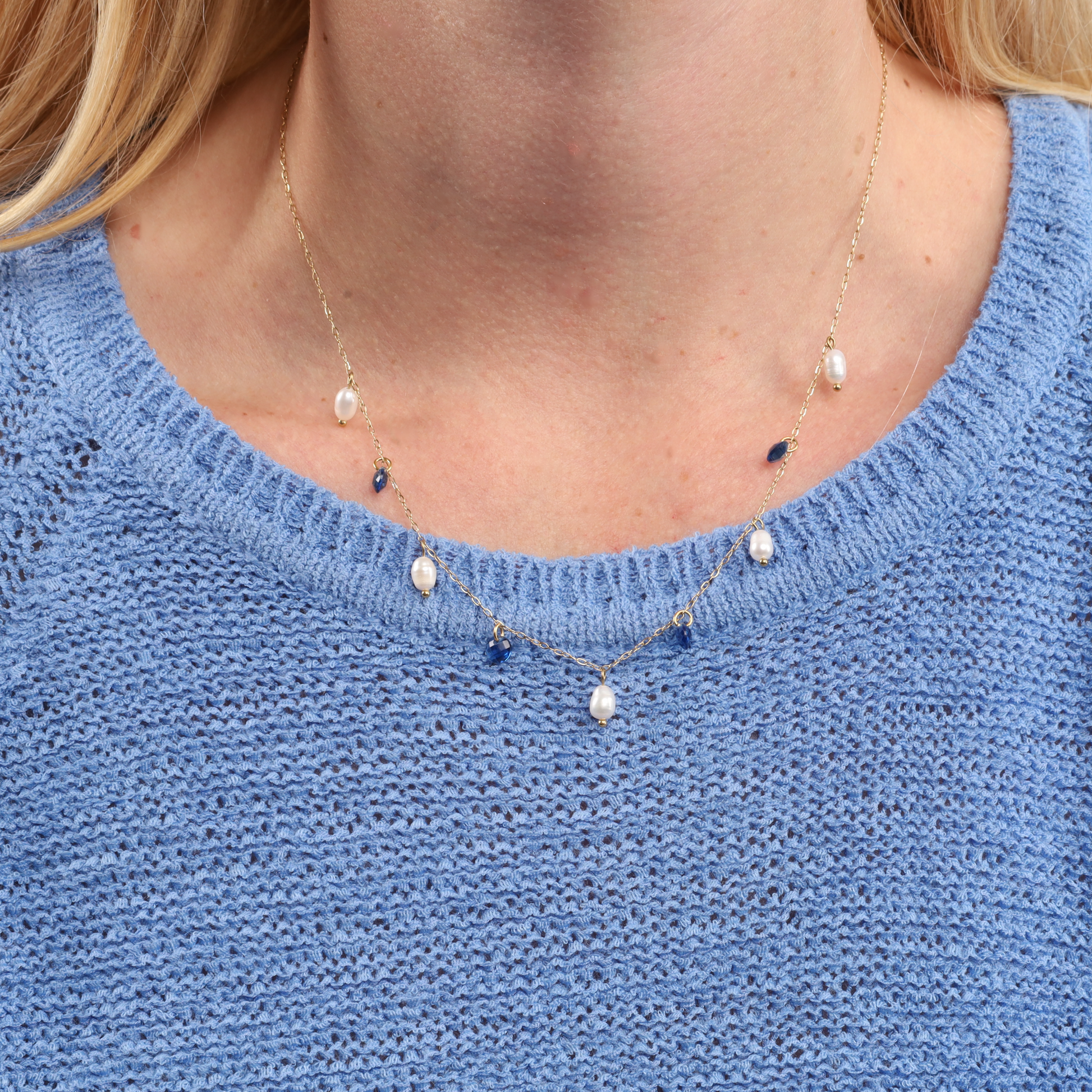 A person wearing a sweater with a Blue Crystal and Pearl Pendant Necklace by Mia Ishaaq®, featuring small pearls and elegant 18k gold plating.