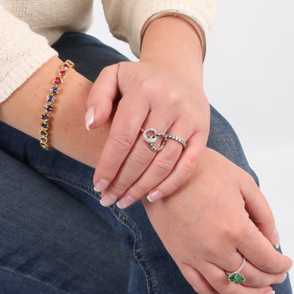 Hands adorned with a jeweled bracelet, a Mia Ishaaq Interlocking Circle Eternity Ring in sterling silver, and an emerald ring resting on blue jeans.