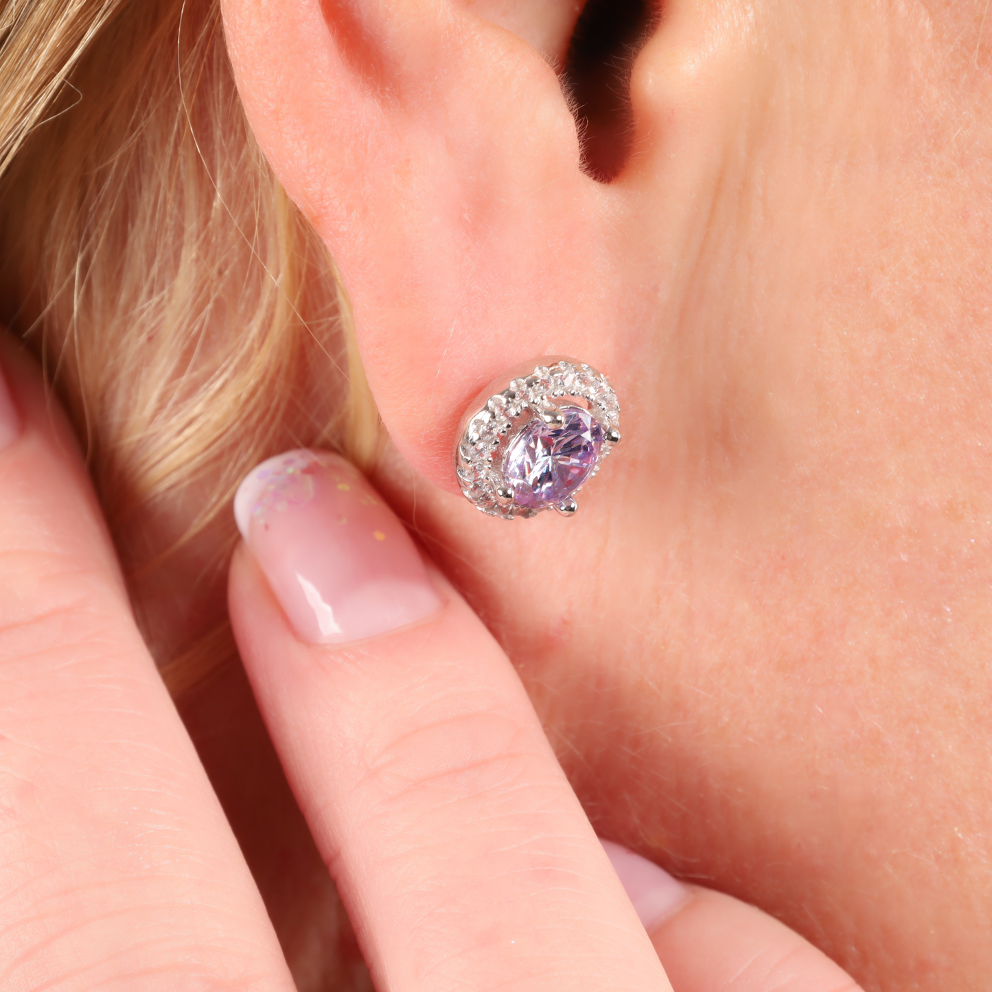 Close-up of an ear adorned with a pair of Mia Ishaaq's 925 Sterling Silver LAVENDER CZ Stud Earrings, with fingers gently touching the earlobe.