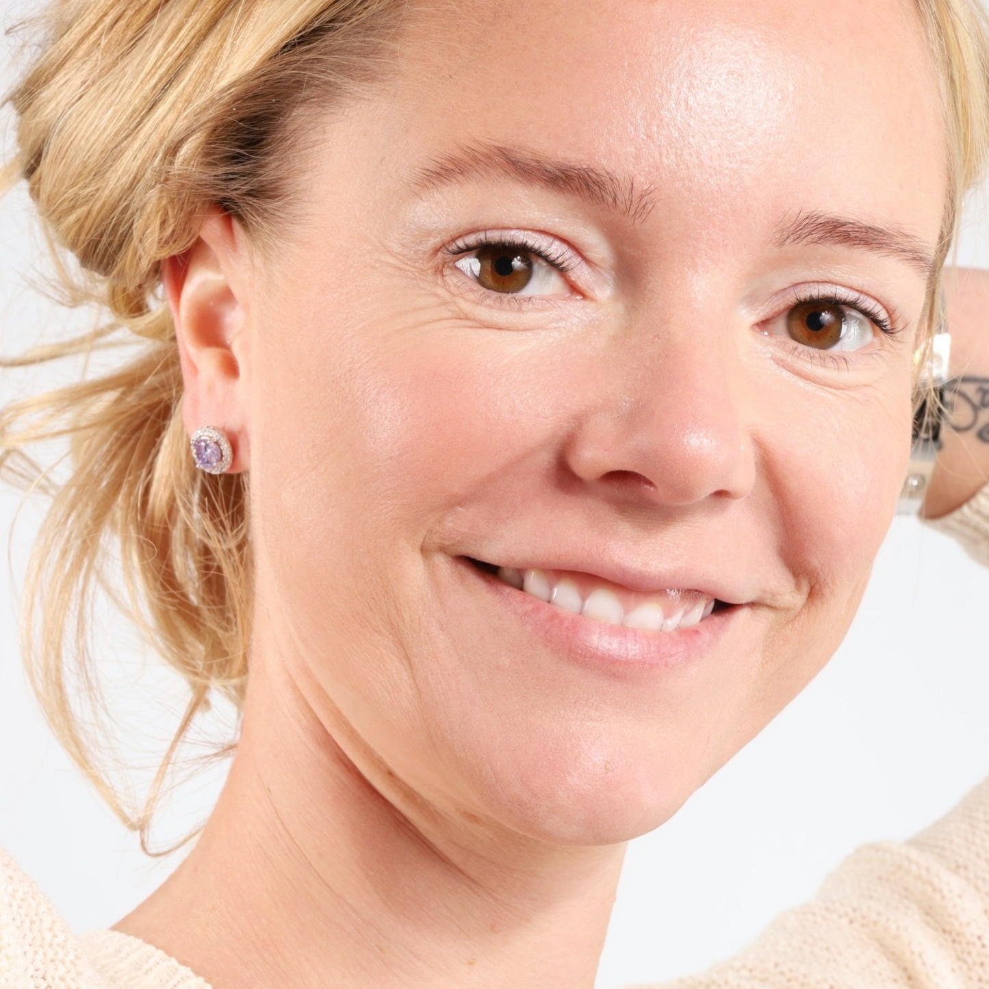 A smiling woman with blonde hair wears elegant 925 Sterling Silver LAVENDER CZ Stud Earrings from Mia Ishaaq, paired with a light-colored sweater against a white background.