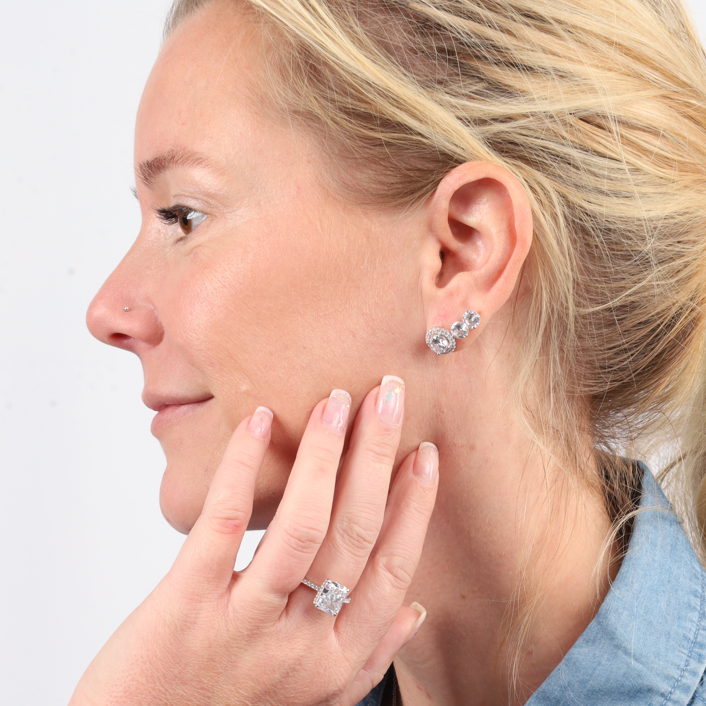 Profile view of a person adorned with Halo Radiance Sparkling Stud Earrings by Mia Ishaaq and a ring, hand gently touching their face, all paired with a denim shirt. The elegant studs capture the light beautifully, adding sophistication to the casual ensemble.