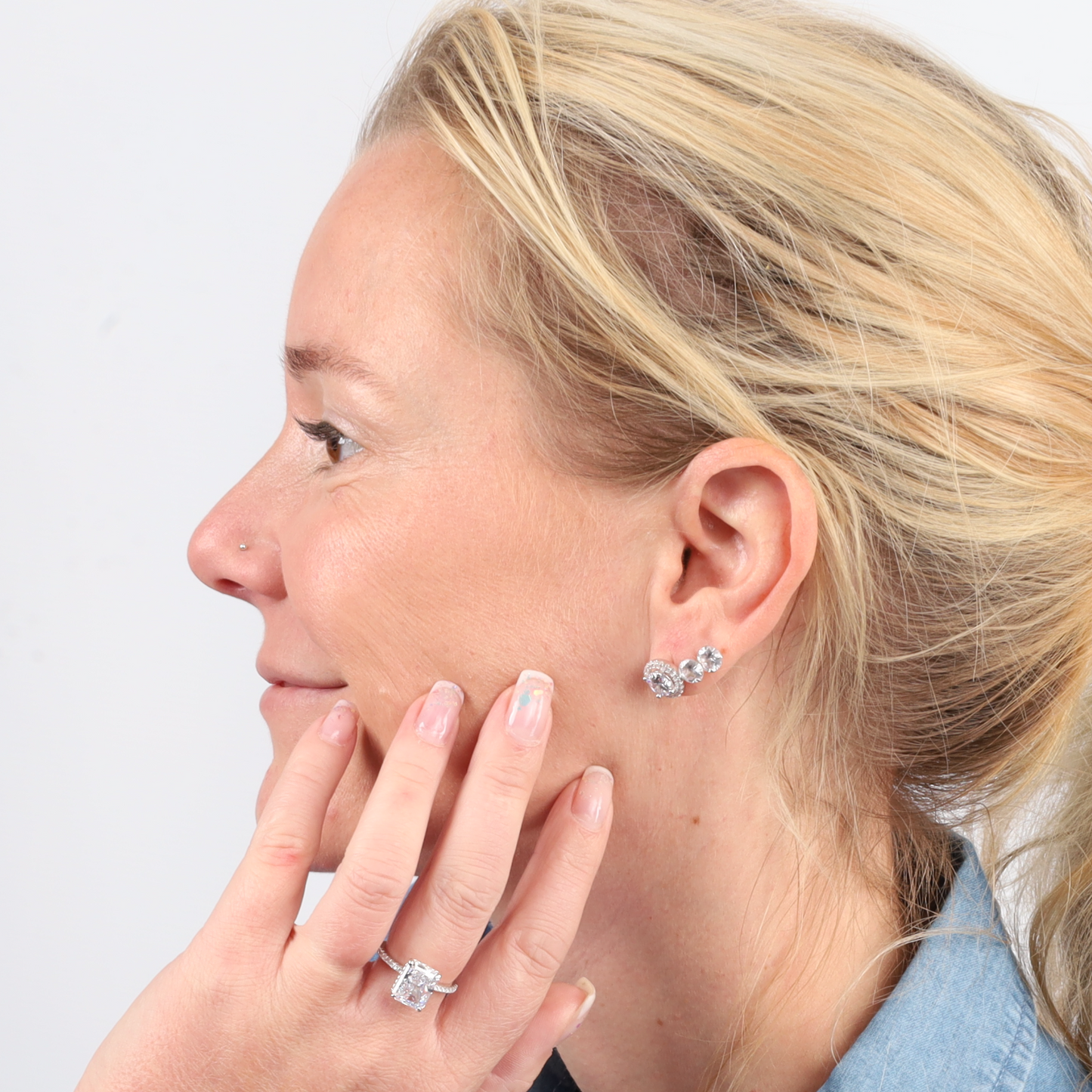 A woman with blonde hair gently touches her face with a smile, adorned in Mia Ishaaq's shimmering cubic zirconia earrings and the exquisite Sterling Silver Clear Radiant Ring.