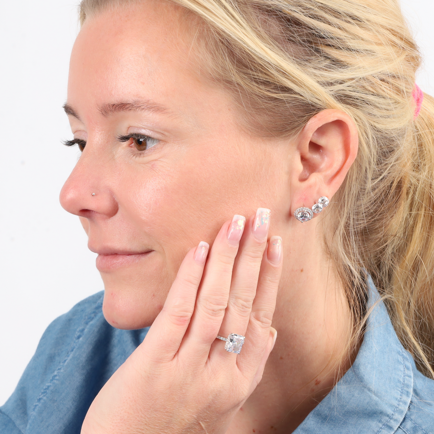 A woman in a denim shirt, showcasing her 925 sterling silver jewelry adorned with Halo Radiance Sparkling Stud Earrings by Mia Ishaaq, smiles slightly.