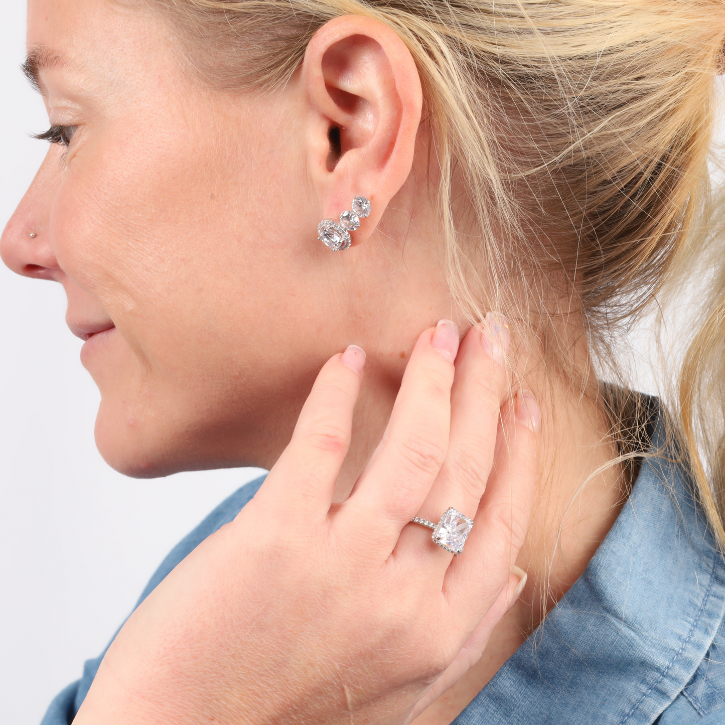 A close-up of a person showcasing their diamond earrings and the Sterling Silver Clear Radiant Ring by Mia Ishaaq, while wearing a denim shirt and gently holding their neck.