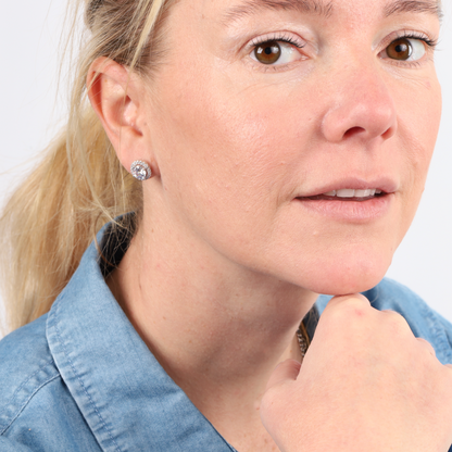 A woman with blonde hair wearing a denim shirt and Mia Ishaaq's Halo Radiance Sparkling Stud Earrings rests her chin on her hand against a white background.