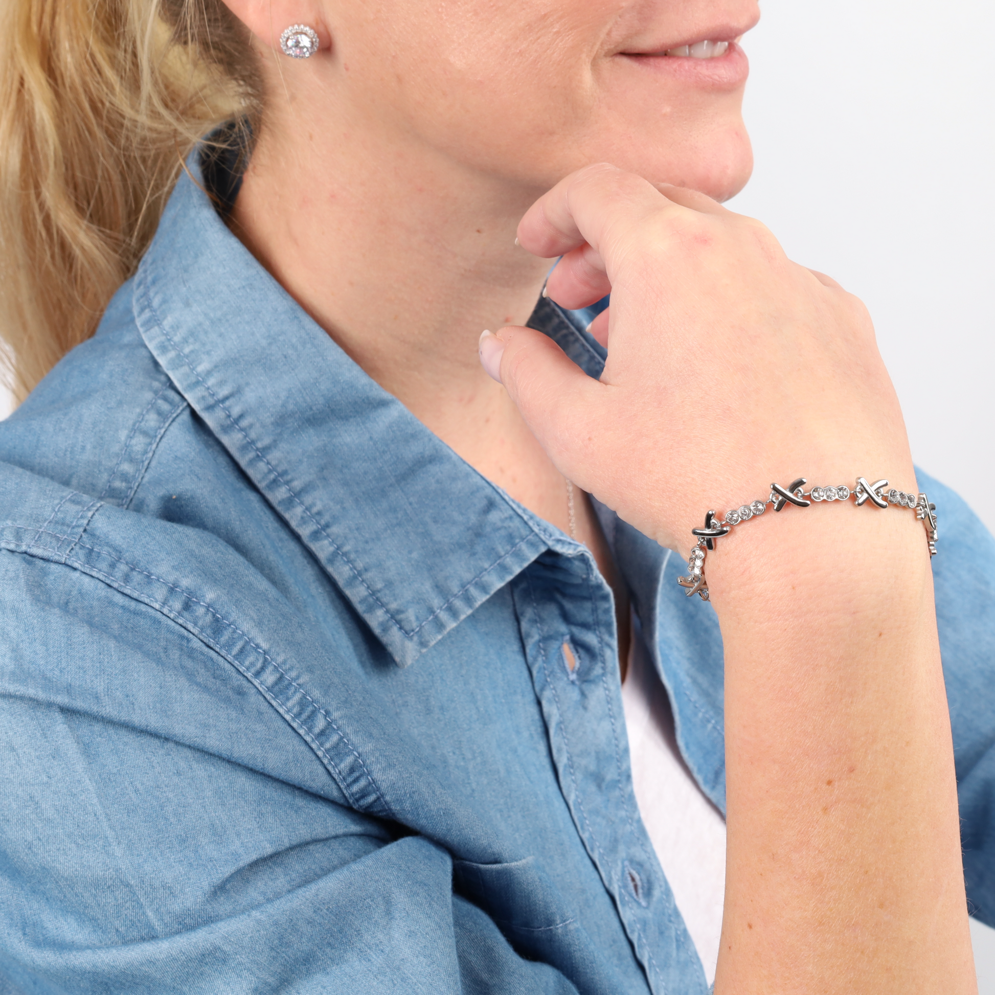 A woman in a denim shirt wearing a Mia Ishaaq Gemma Owen Inspired XO Cross Bracelet and diamond stud earrings, with her hand gently resting on her chin.