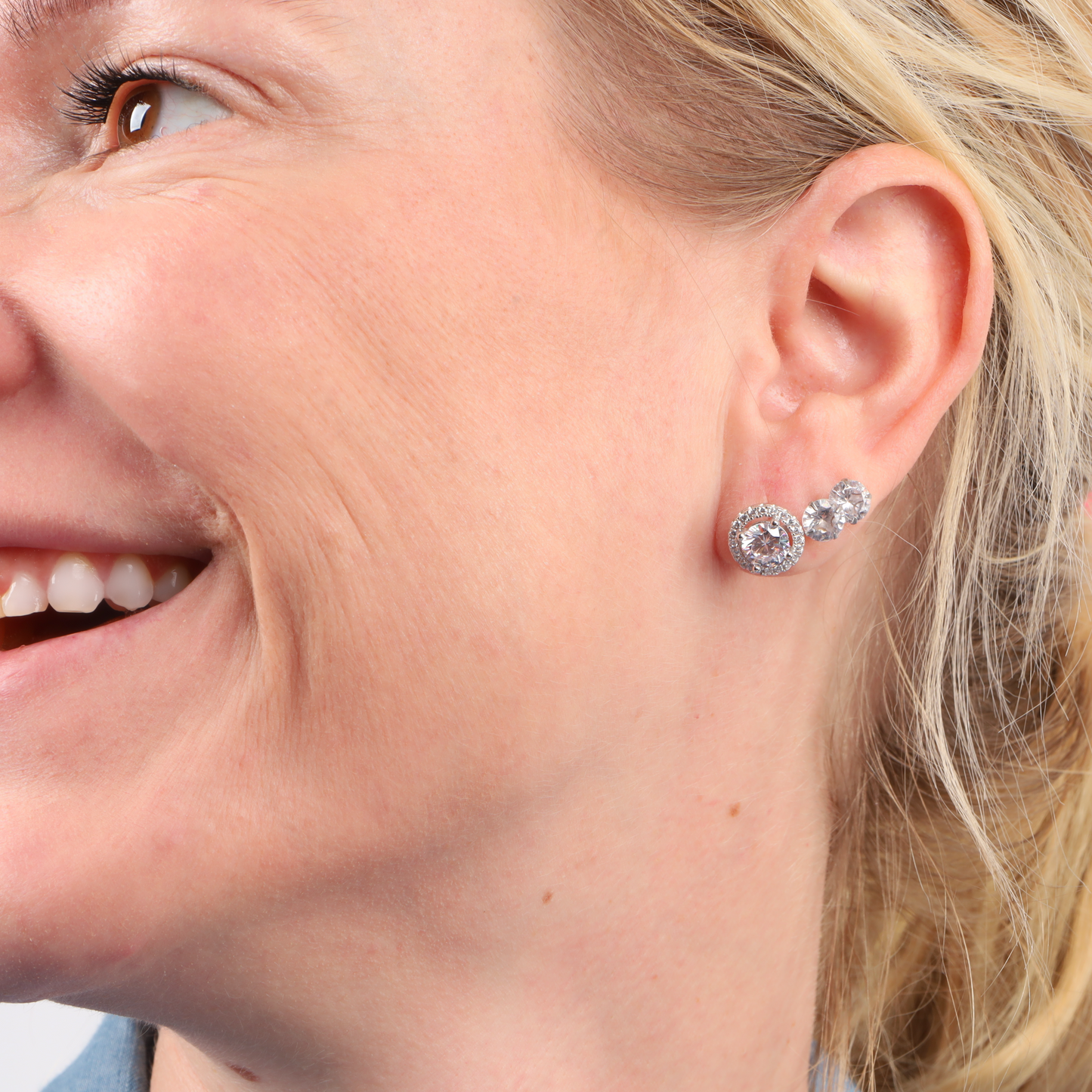 Close-up of a smiling person wearing Halo Radiance Sparkling Stud Earrings by Mia Ishaaq, featuring clear CZ studs set in 925 sterling silver.