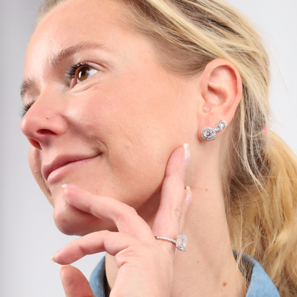 A person smiling, touching their face while wearing the Halo Radiance Sparkling Stud Earrings by Mia Ishaaq and a sparkling ring made of 925 sterling silver, with their hair tied back.