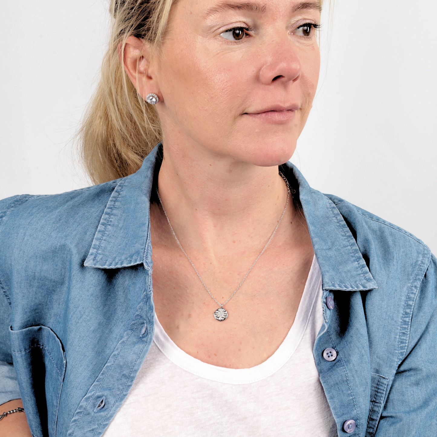 Woman in a denim shirt wearing the Polaris Coin Pendant Necklace by Mia Ishaaq® in sterling silver, facing left, with a neutral expression and earrings.