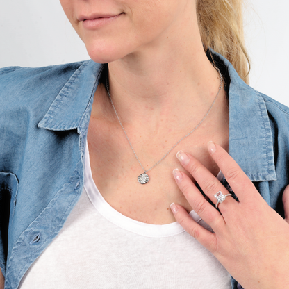 A person wearing a denim shirt showcases the Polaris Coin Pendant Necklace in silver from Mia Ishaaq®, highlighted by its circular charm, and pairs it with a ring featuring a striking gemstone.