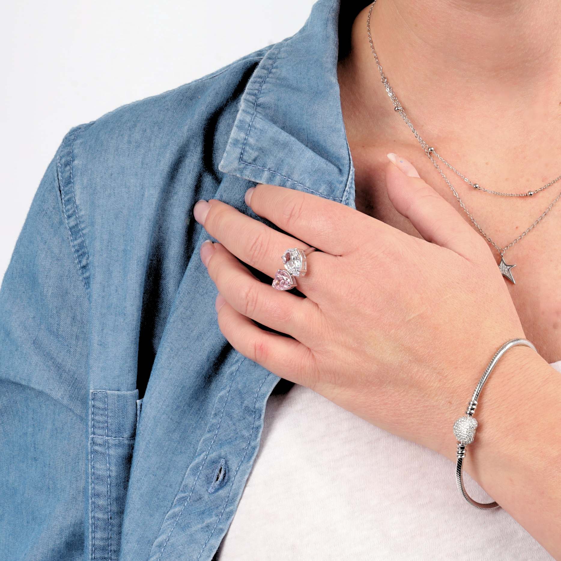 A person dressed in a denim shirt and accessorized with a sleek bracelet and star charm necklace touches their chest, where the Mia Ishaaq Sterling Silver Radiant Duo Ring - adjustable - sparkles with brilliant cubic zirconia stones.