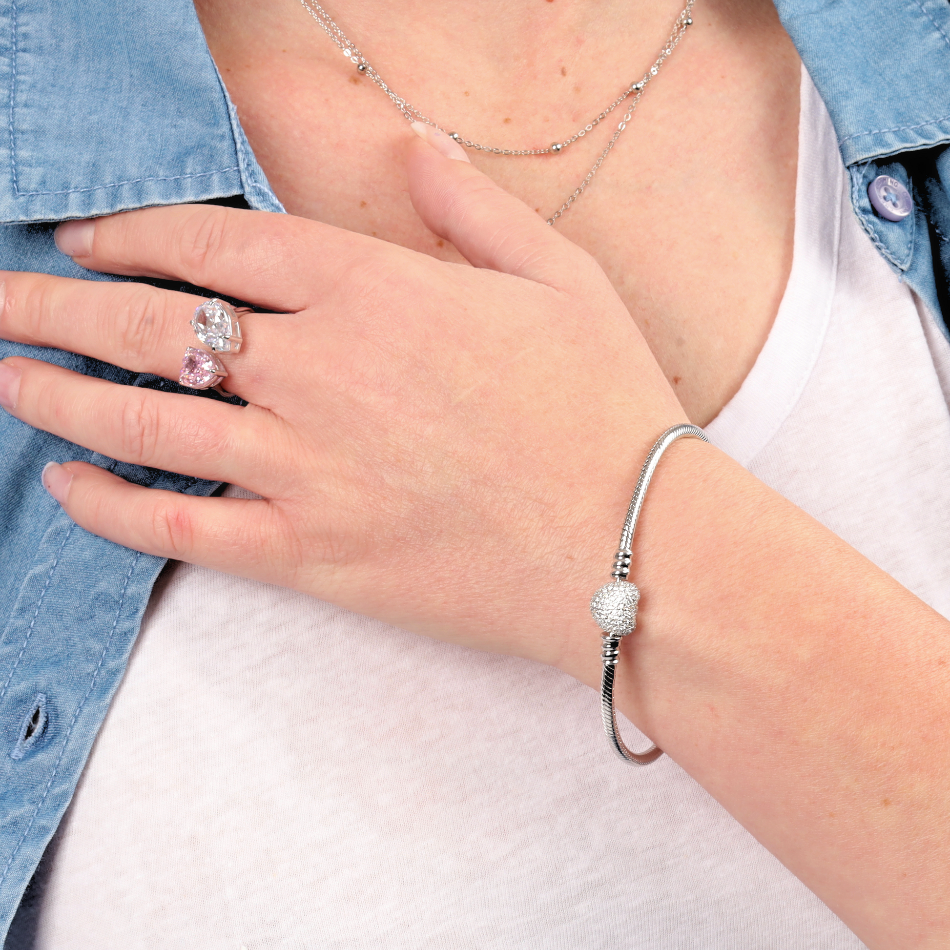 A person wearing the ATHENAIE Pave Heart Snake Chain Charm Bracelet and ring, with their hand resting on their chest. They are dressed in a white top and denim shirt, subtly highlighting the elegance of the jewelry.