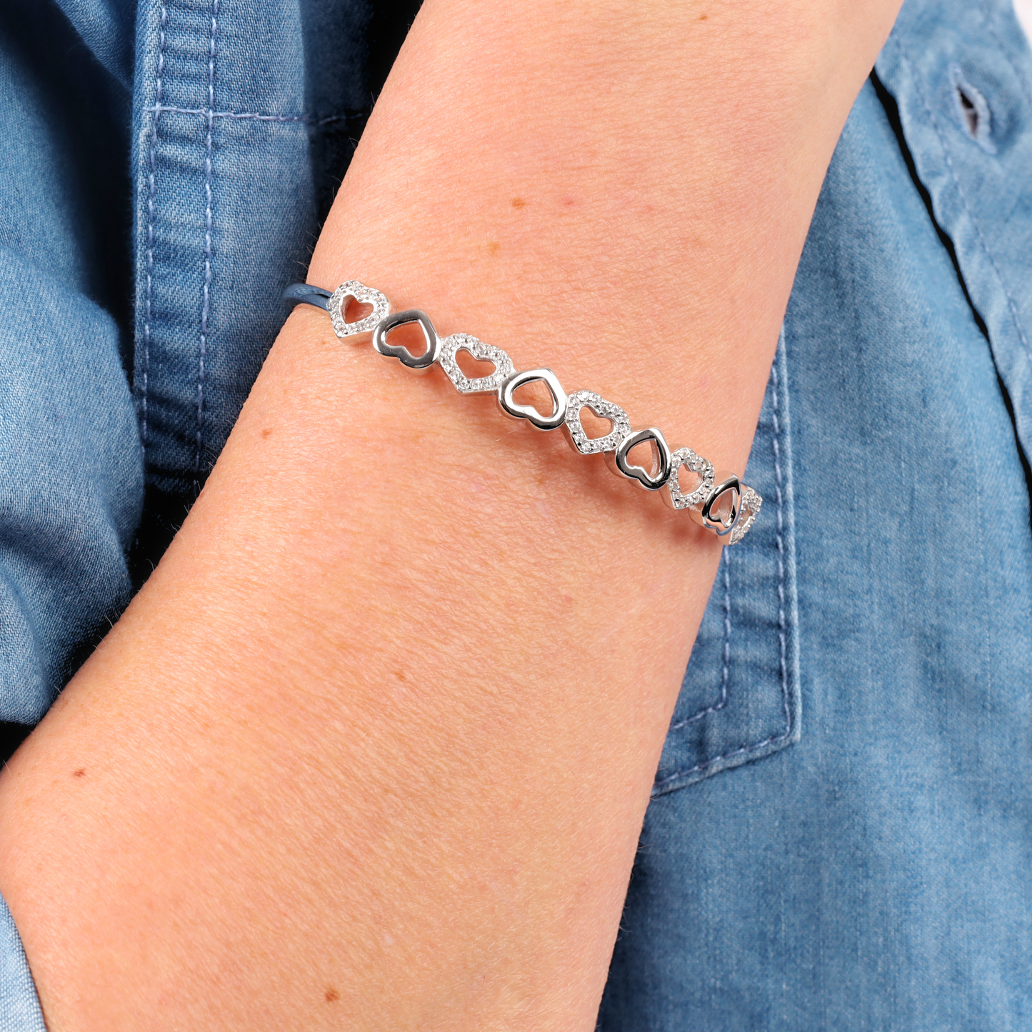 A person elegantly rests a Mia Ishaaq Sterling Silver Heart Bangle Bracelet on denim shirt fabric.