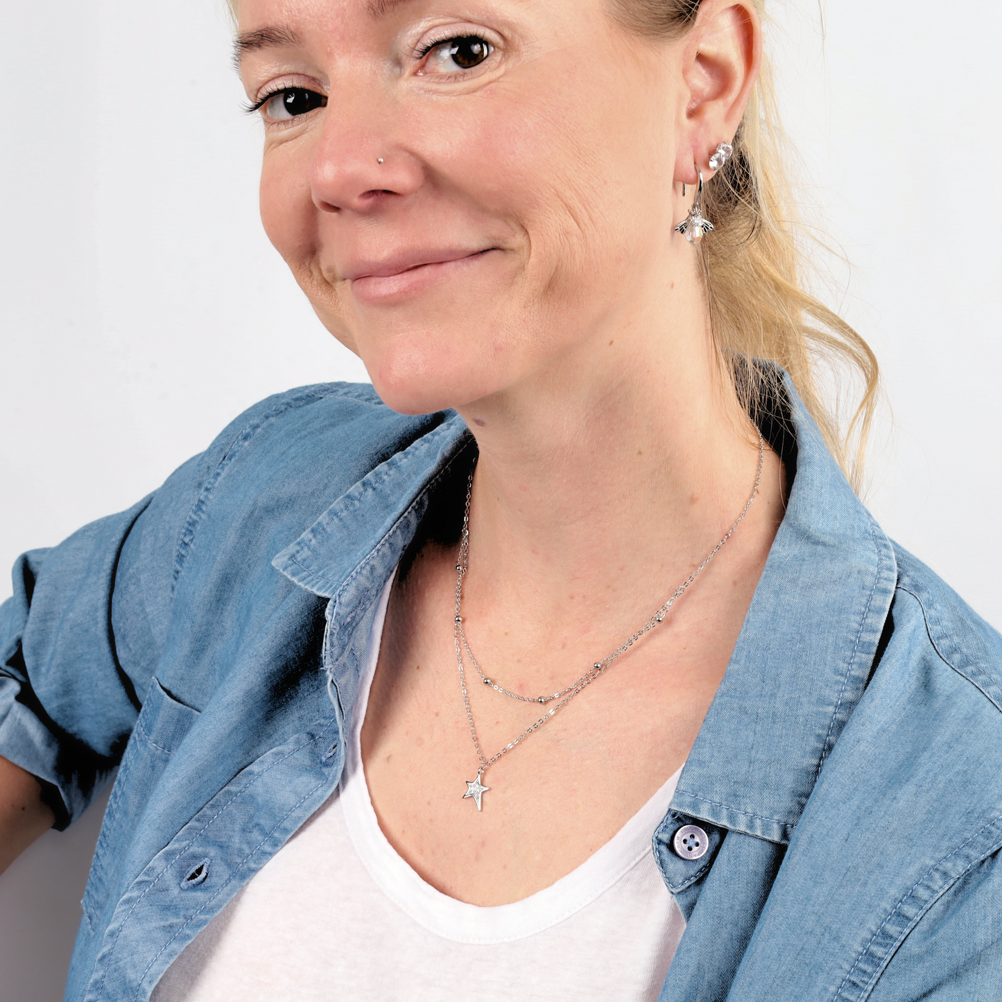 A woman in a denim shirt wears Mia Ishaaq's Crystal Queen Bee Dangle Earrings and necklaces, smiling at the camera.