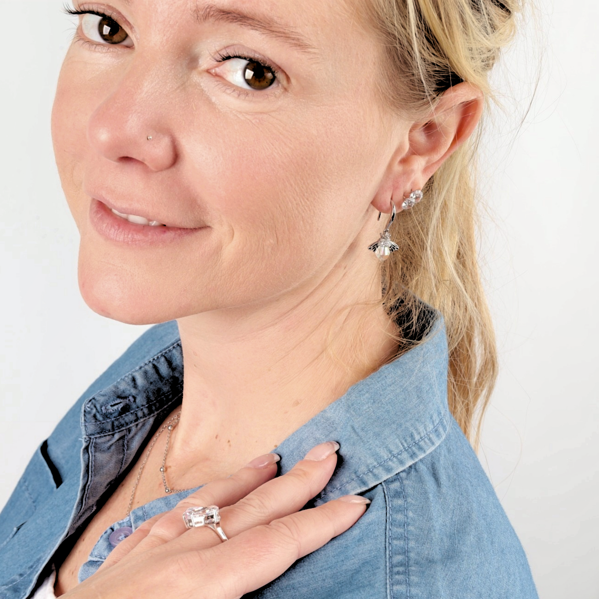 A smiling woman with a nose piercing, wearing Mia Ishaaq's Crystal Queen Bee Dangle Earrings and a ring, in a blue shirt, looking over her shoulder.