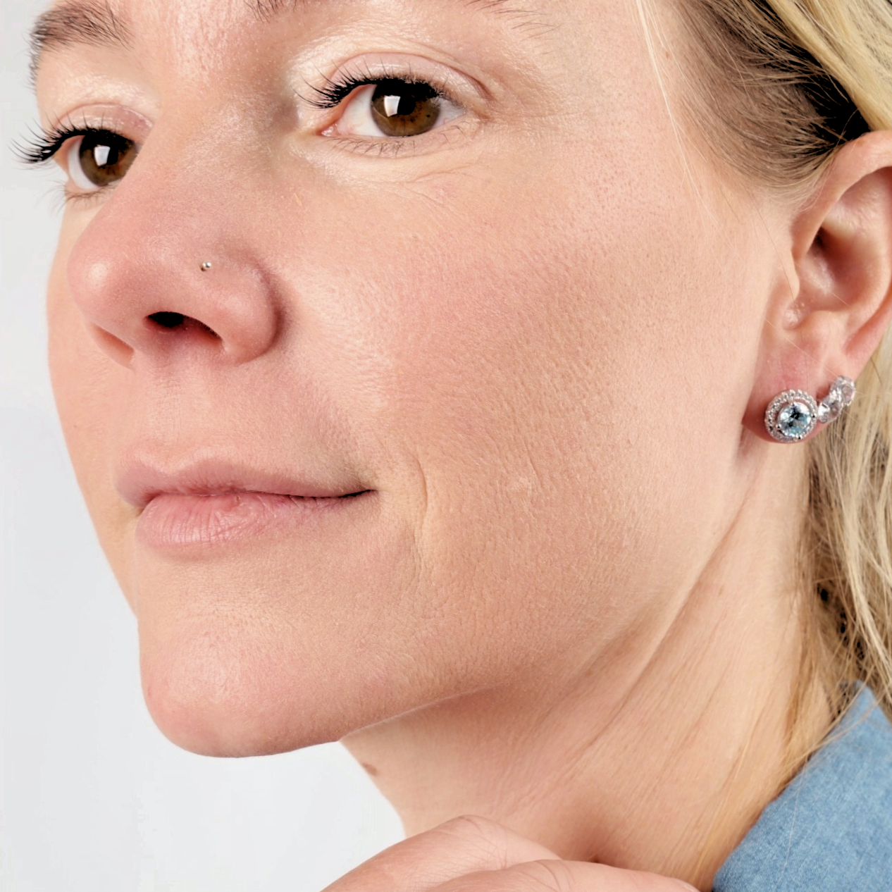 A close-up of a woman wearing Mia Ishaaq's 925 Sterling Silver AQUAMARINE CZ Stud Earrings and a nose stud, paired with a light blue top.