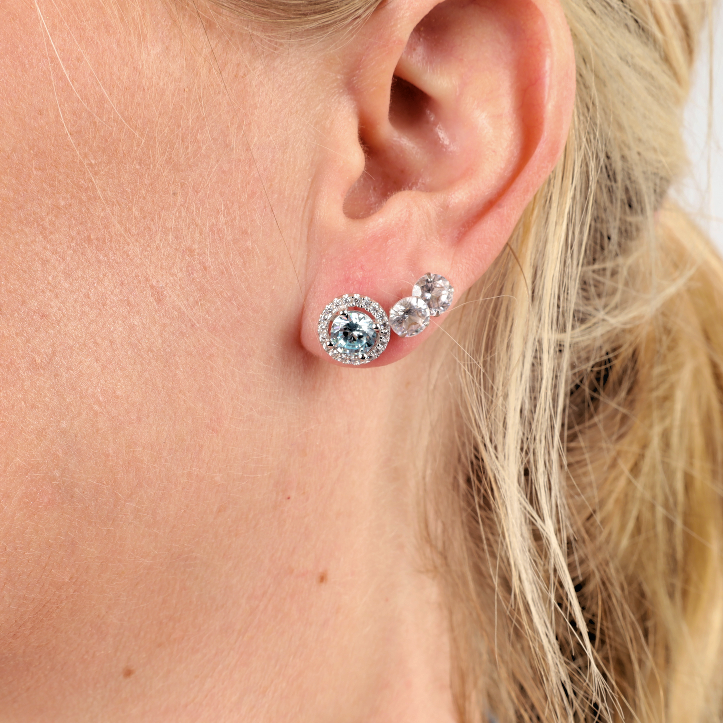 Close-up of a woman’s ear adorned with Mia Ishaaq 925 Sterling Silver AQUAMARINE CZ Stud Earrings, featuring an Aquamarine CZ gemstone and a small clear gem.