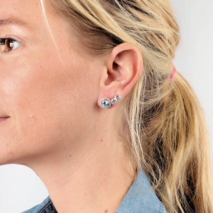 A woman with blonde hair wears three pairs of Mia Ishaaq's 925 Sterling Silver AQUAMARINE CZ Stud Earrings, each featuring a sparkling Aquamarine CZ stone, against a light background.