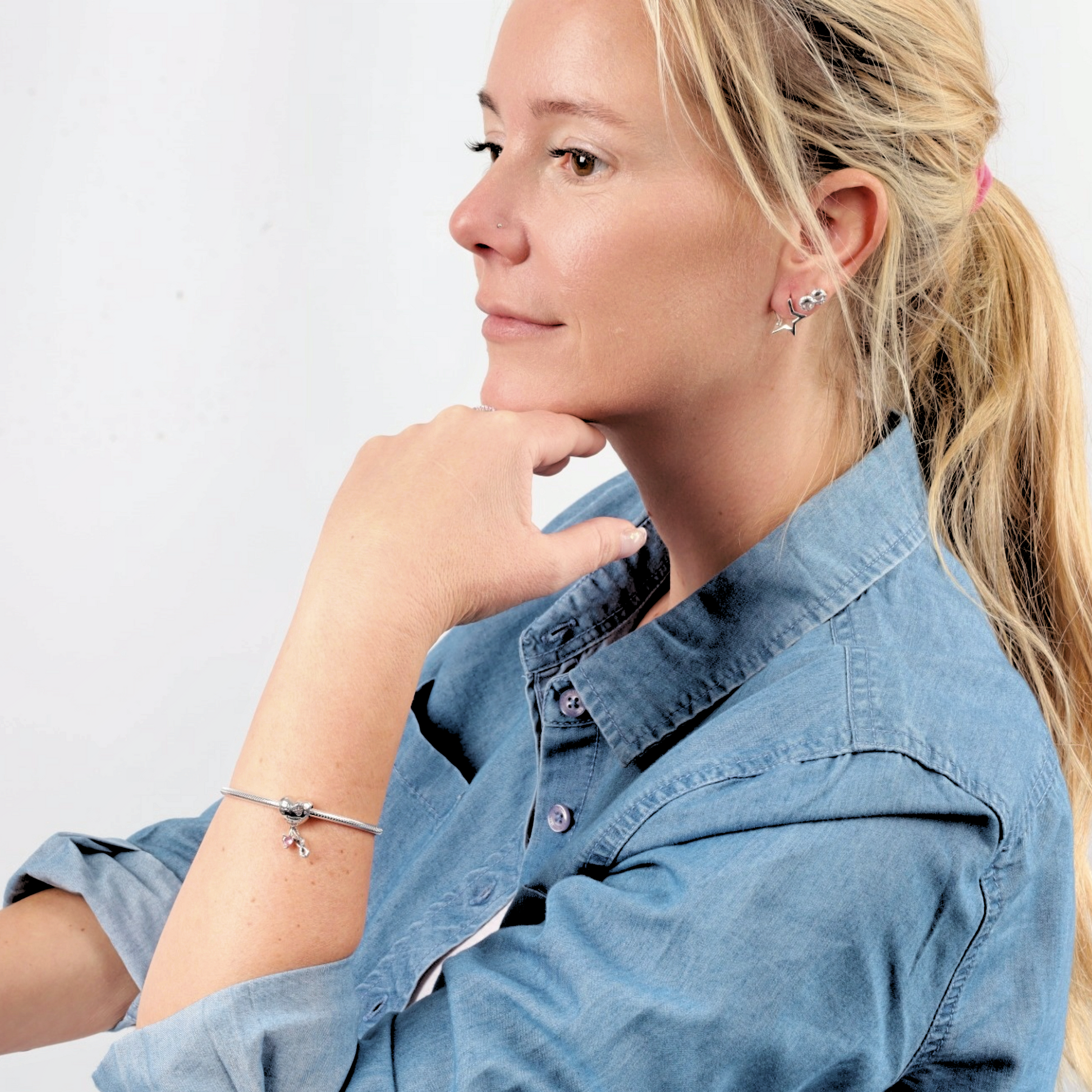 A woman in a denim shirt gazes thoughtfully, showcasing a Snake Chain Charm Bracelet by Mia Ishaaq on her wrist, set against a white background.