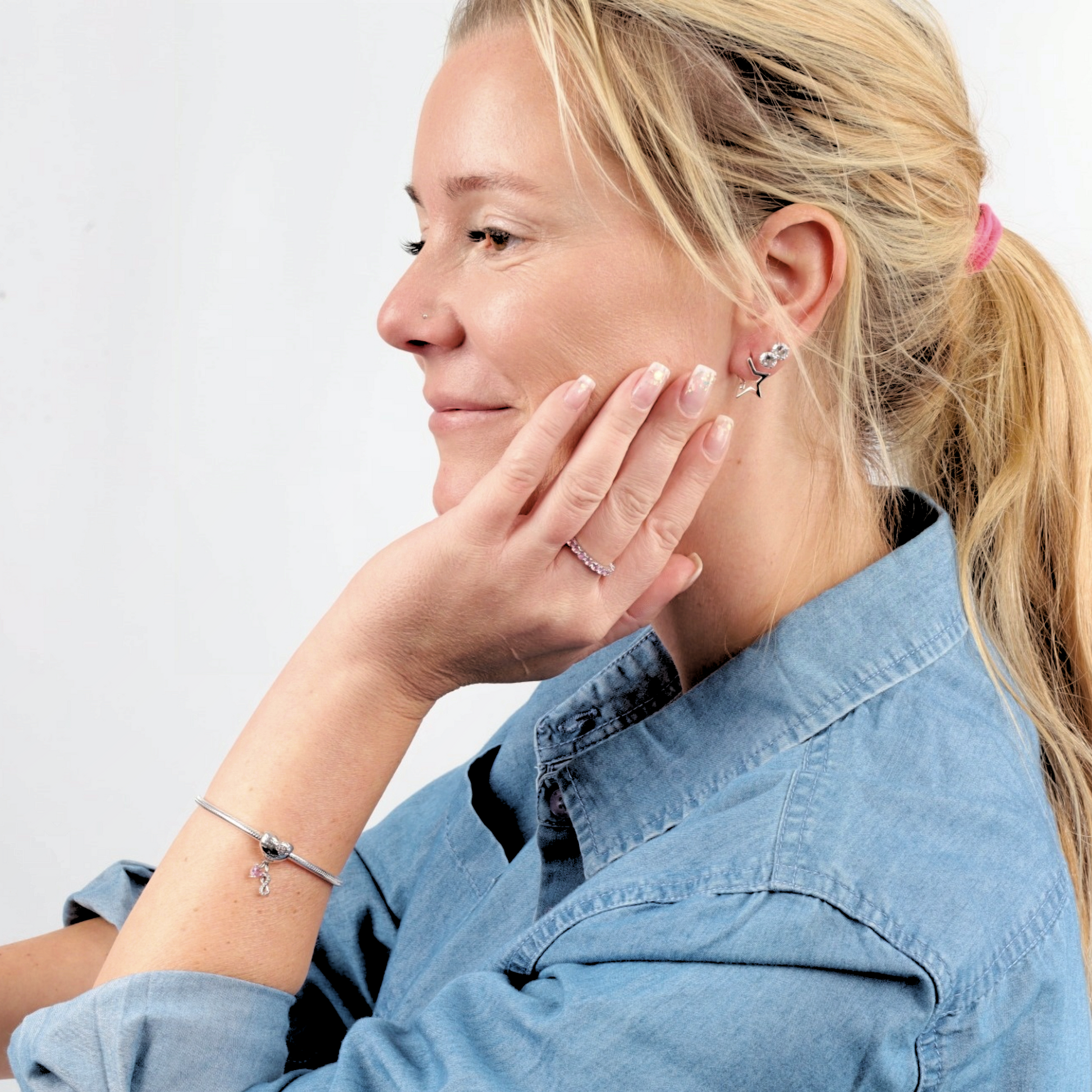 A woman in a denim shirt, shown from the side, wears sterling silver jewelry and a snake chain charm bracelet by Mia Ishaaq, with her hand held near her face.