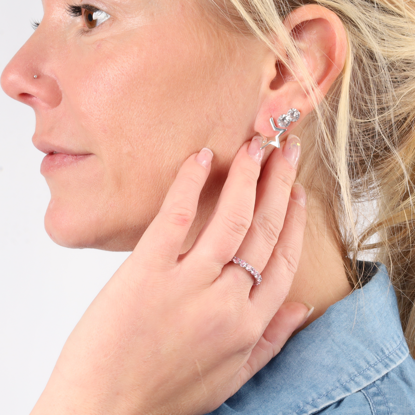 A person wearing a star-shaped earring and a jeweled ring, captured in a close-up side profile view, exemplifies elegance. The Sterling Silver Pink Ring Band from Mia Ishaaq adds a touch of sparkle, enhancing the overall allure.