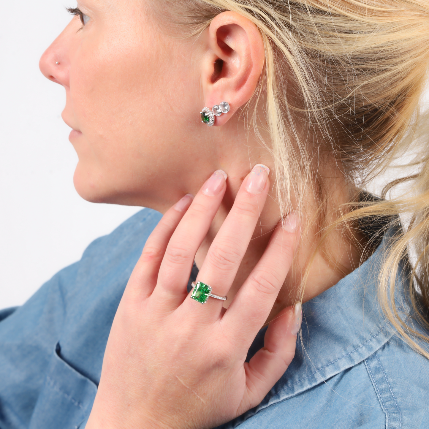 A person with blonde hair is wearing nature-inspired jewelry, including the Emerald Glow Radiant Studs by Mia Ishaaq, a green gemstone ring, and matching pieces, all paired with a blue shirt.