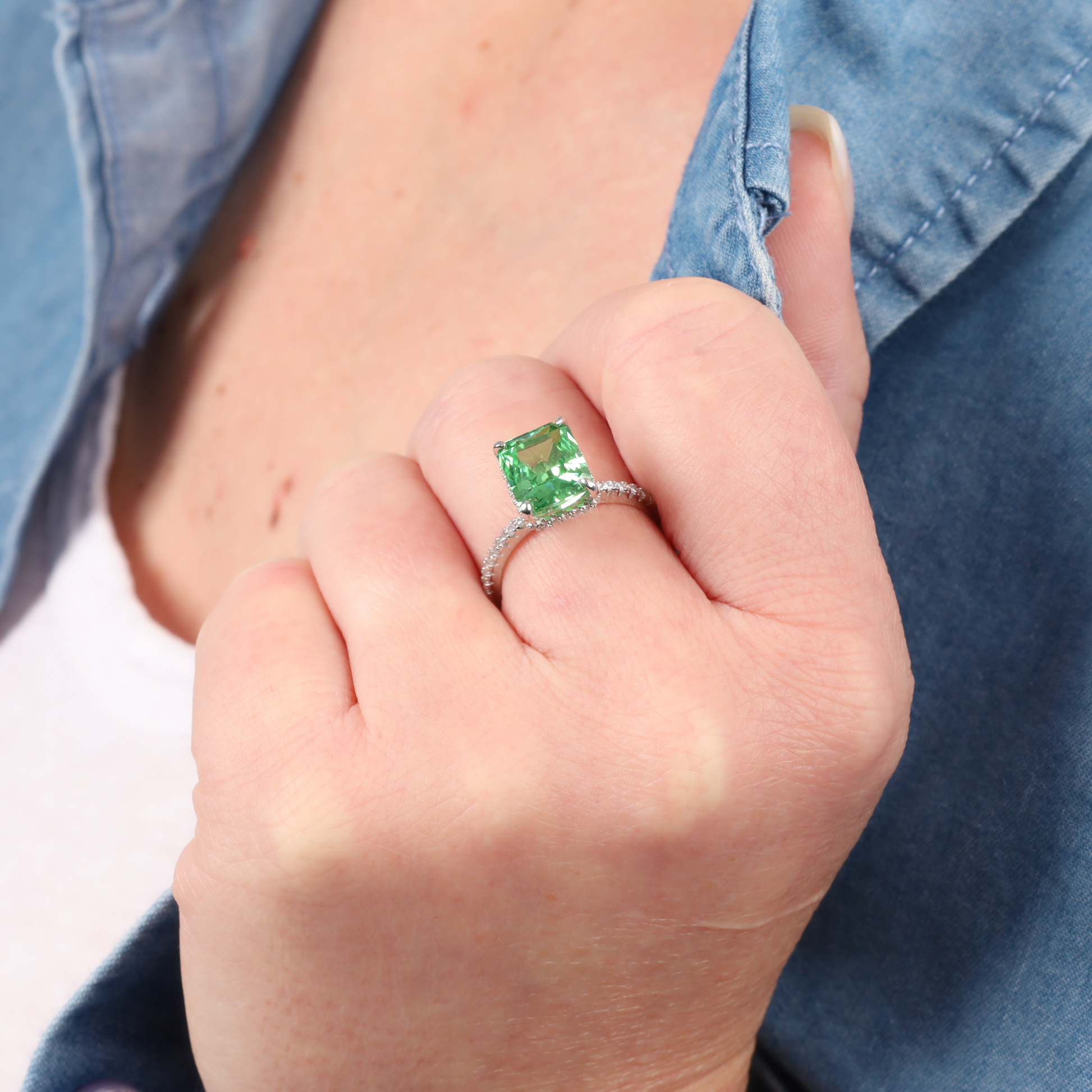 Close-up of a hand adorned with the Crushed Ice Green Radiant Promise Ring by Mia Ishaaq, featuring a large green cubic zirconia stone, set against the backdrop of a blue denim shirt.