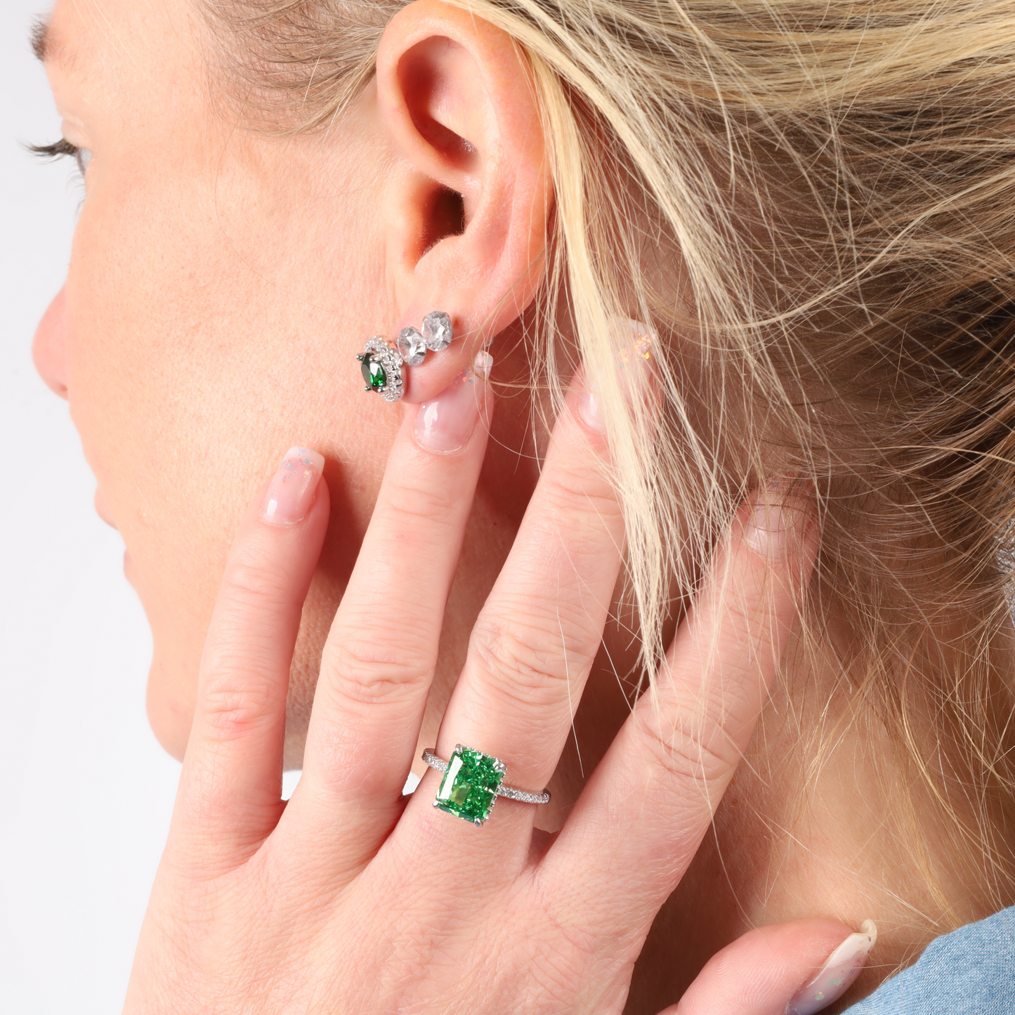 A woman adorned with the Crushed Ice Green Radiant Promise Ring by Mia Ishaaq complements her gemstone earrings as she gently touches her ear.