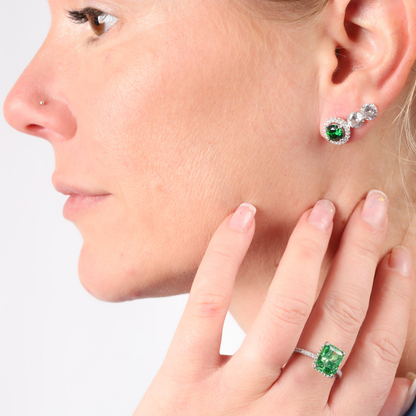 Person wearing green gemstone earrings and a Crushed Ice Green Radiant Promise Ring by Mia Ishaaq, touching face with their right hand against a white background.