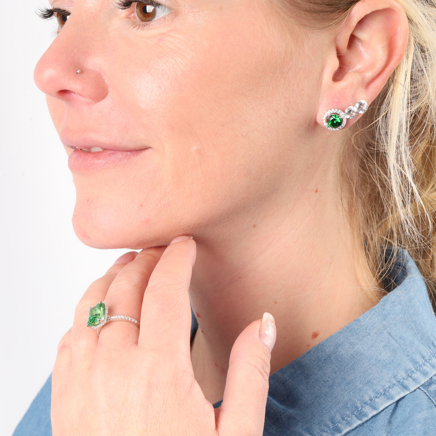 A woman adorned with elegant Emerald Glow Radiant Studs from Mia Ishaaq and a matching ring glances to the side, her blue shirt providing a perfect backdrop for the nature-inspired jewelry.