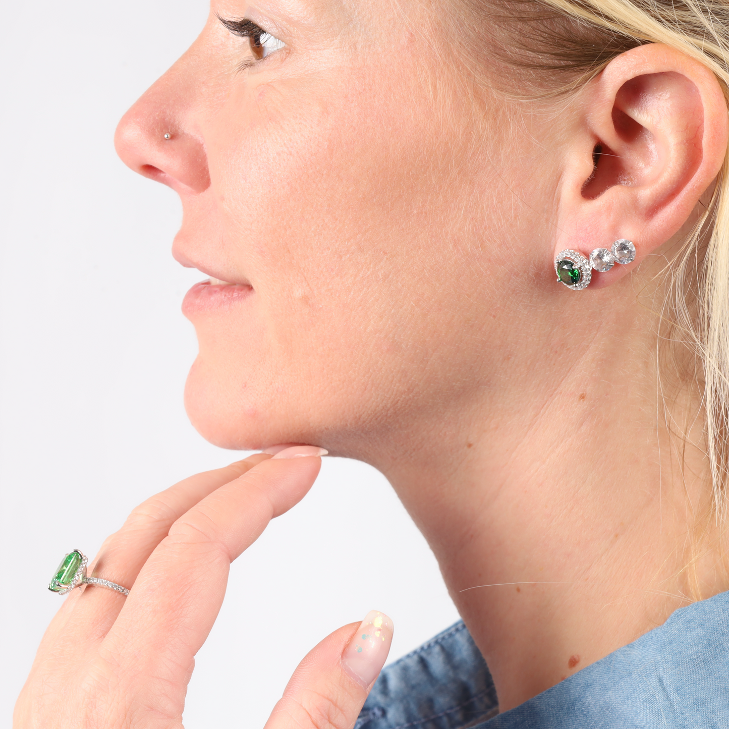 Person with green cubic zirconia stone earrings and a Crushed Ice Green Radiant Promise Ring by Mia Ishaaq, wearing a blue top, looking to the side.