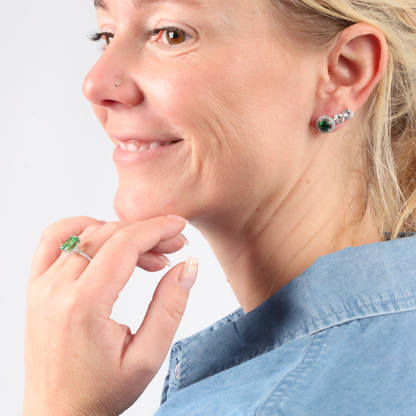 A woman, adorned with nature-inspired jewellery, smiles in a denim shirt, showcasing her Emerald Glow Radiant Studs by Mia Ishaaq and matching ring.